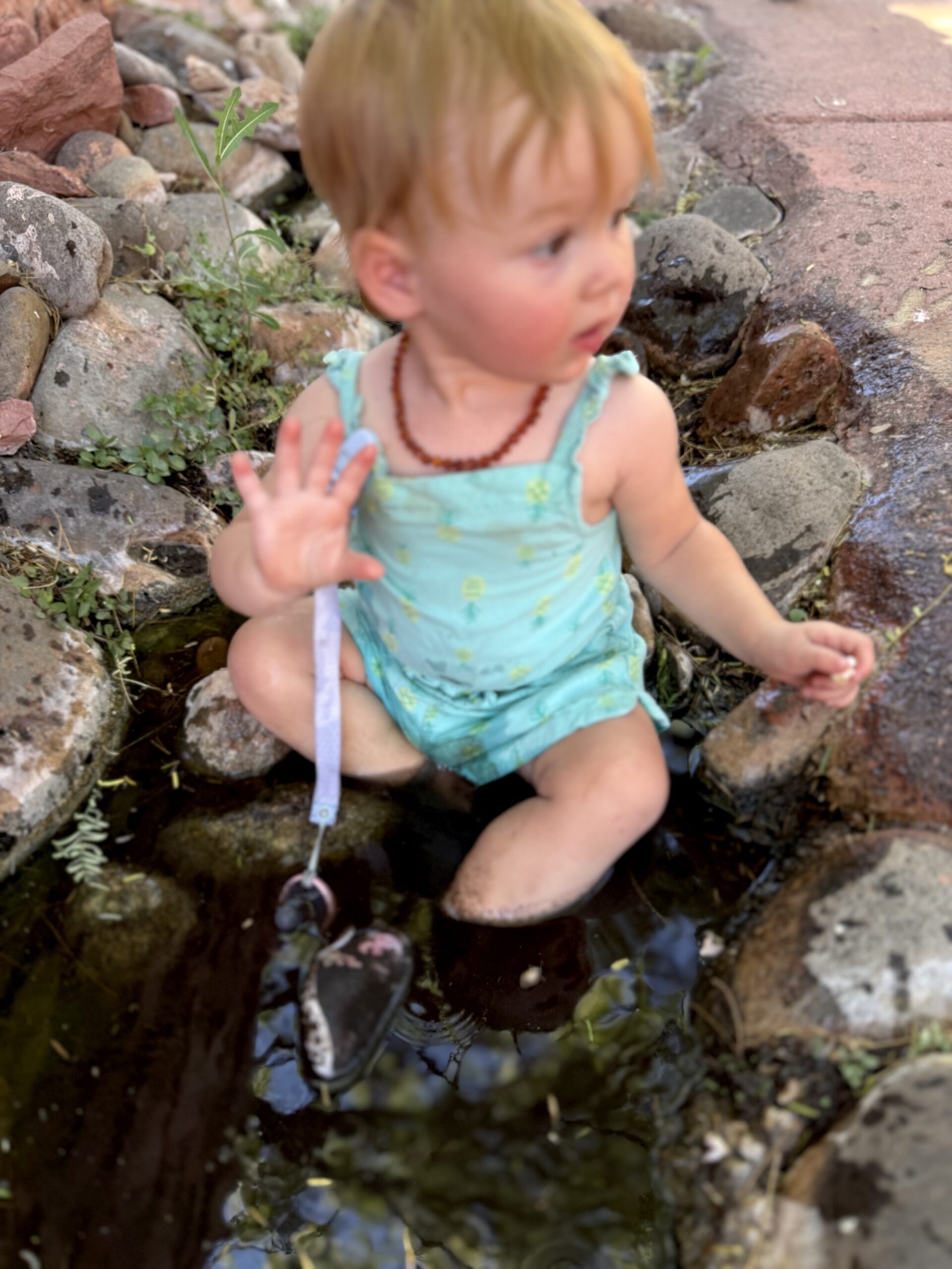 toddler sitting in muddy creek bank at Oak Creek Sedona, hands deep in red mud, yellow dress — letting children play in dirt